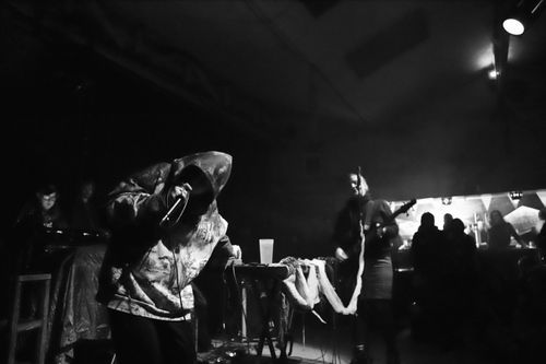 Photo en noir et blanc, on voit une personne en sweat à capuche brandissant un micro, son visage est invisible. une bassiste au fond elle aussi devant un micro. Au dernier plan on devine un bar et des spectateurs.